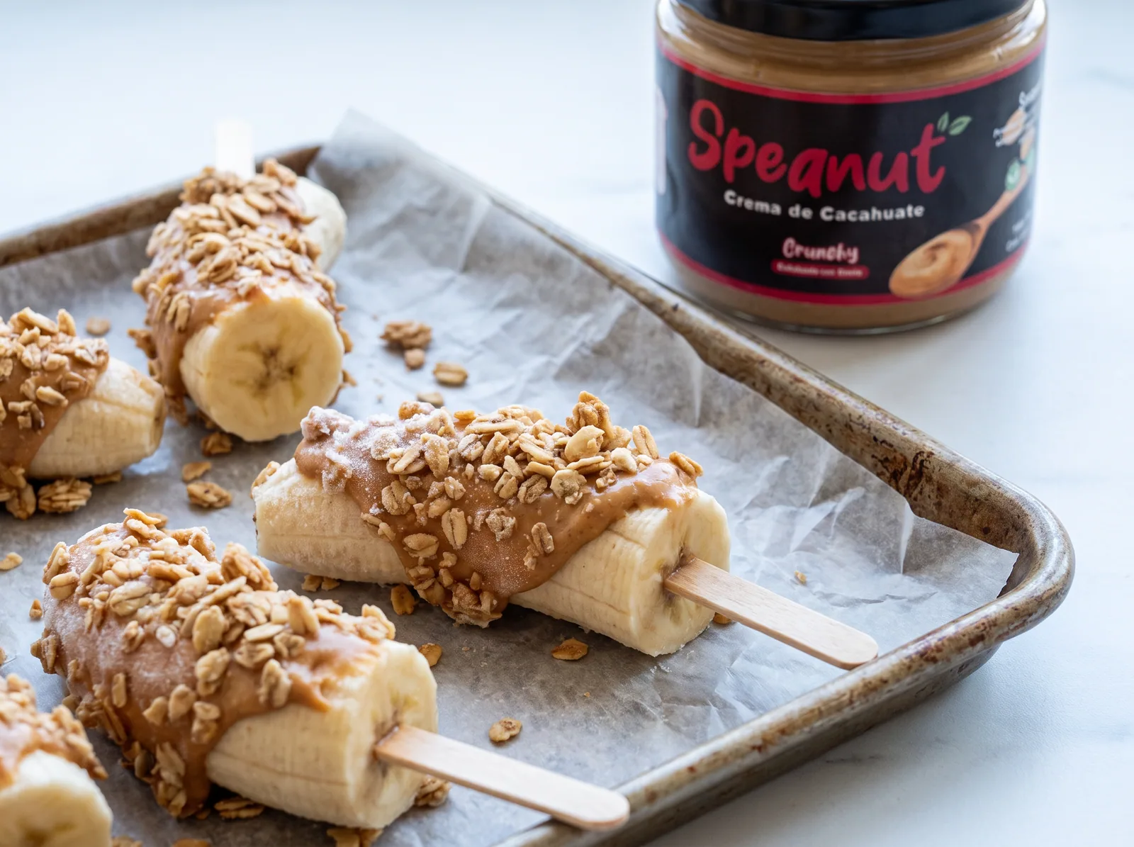 Plátanos en palito cubiertos de crema de cacahuate y granola, sobre una charola con papel encerado