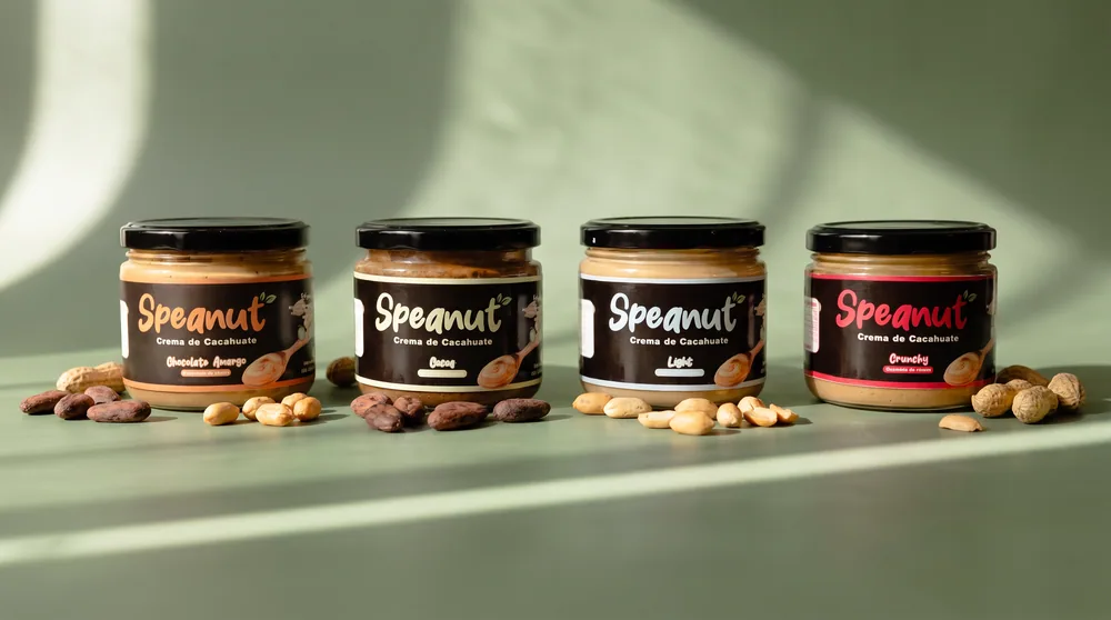 Las cuatro presentaciones de Speanut alineadas sobre fondo verde salvia con cacahuates y granos de cacao